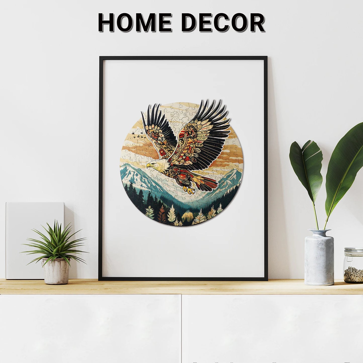 A framed Benlarkytoys 200pcs Tribal Eagle Wooden Jigsaw Puzzle for Adults artwork hangs above a light wood cabinet with a potted plant, cactus, and green leaves. The words HOME DECOR are displayed above.