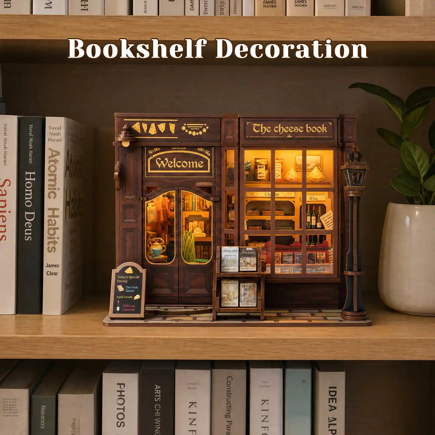 A miniature bookshelf decoration shaped like a cozy bookshop storefront sits on a wooden shelf, surrounded by books and a potted plant. The shop features warm yellow interior lighting, decorative windows, a 'Welcome' sign, and a small streetlamp, with the text 'Bookshelf Decoration' overlaid at the top.