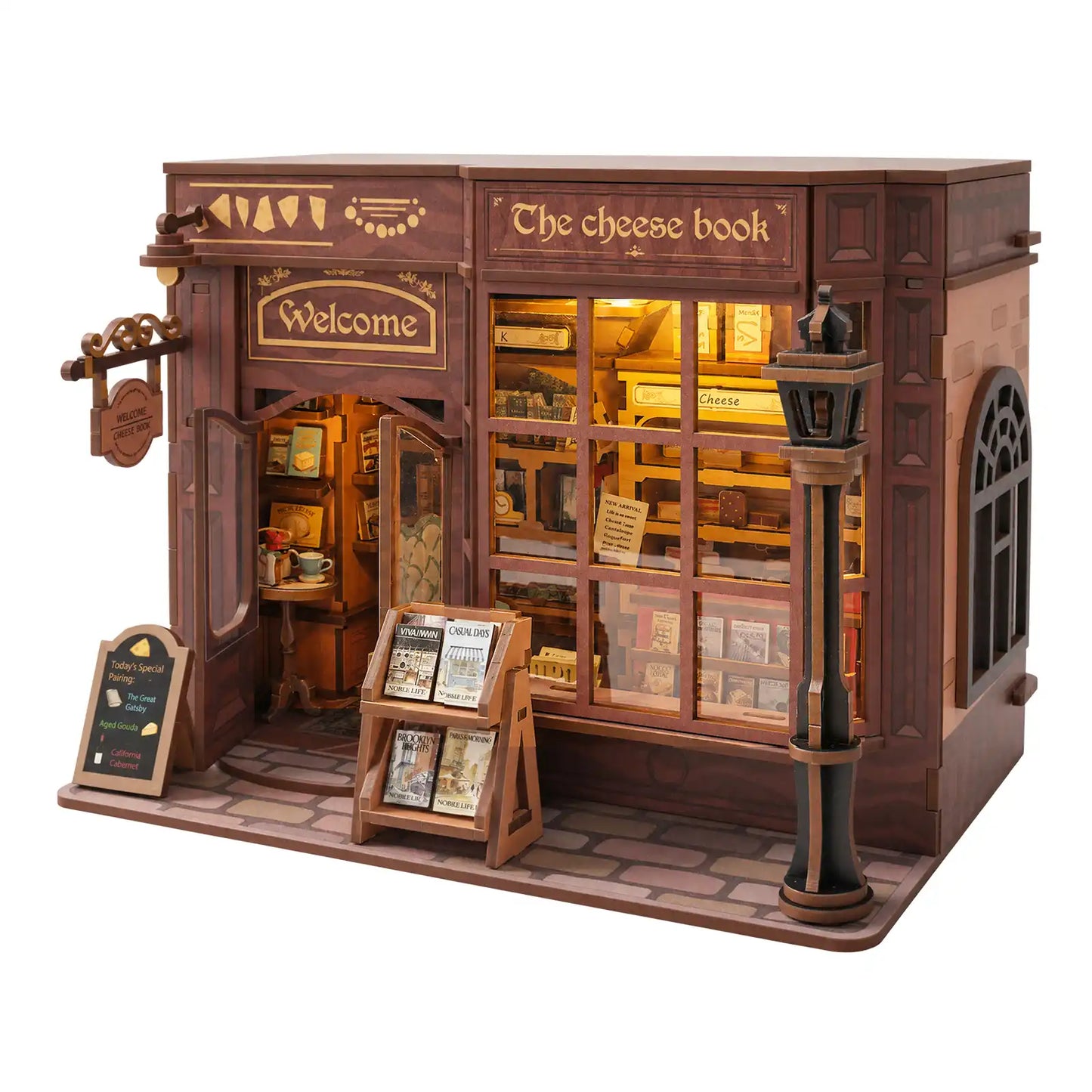 A wooden model of a quaint shop called 'The cheese book' on a clean white background. The miniature building features dark brown wood paneling, warm yellow glowing windows, a street lamp, a sidewalk, and a small sign on the left.