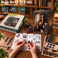 A DIY book nook kit shown on a wooden table with a person's hands assembling pieces from an instruction booklet. The kit includes a decorative wooden nook structure with a lit arched window, the product box, loose components, and accessories.