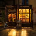 A miniature wooden model of a cozy shop front with warm glowing windows, labeled 'Welcome' and 'The cheese book,' displayed on a reflective surface against a softly blurred dark background.