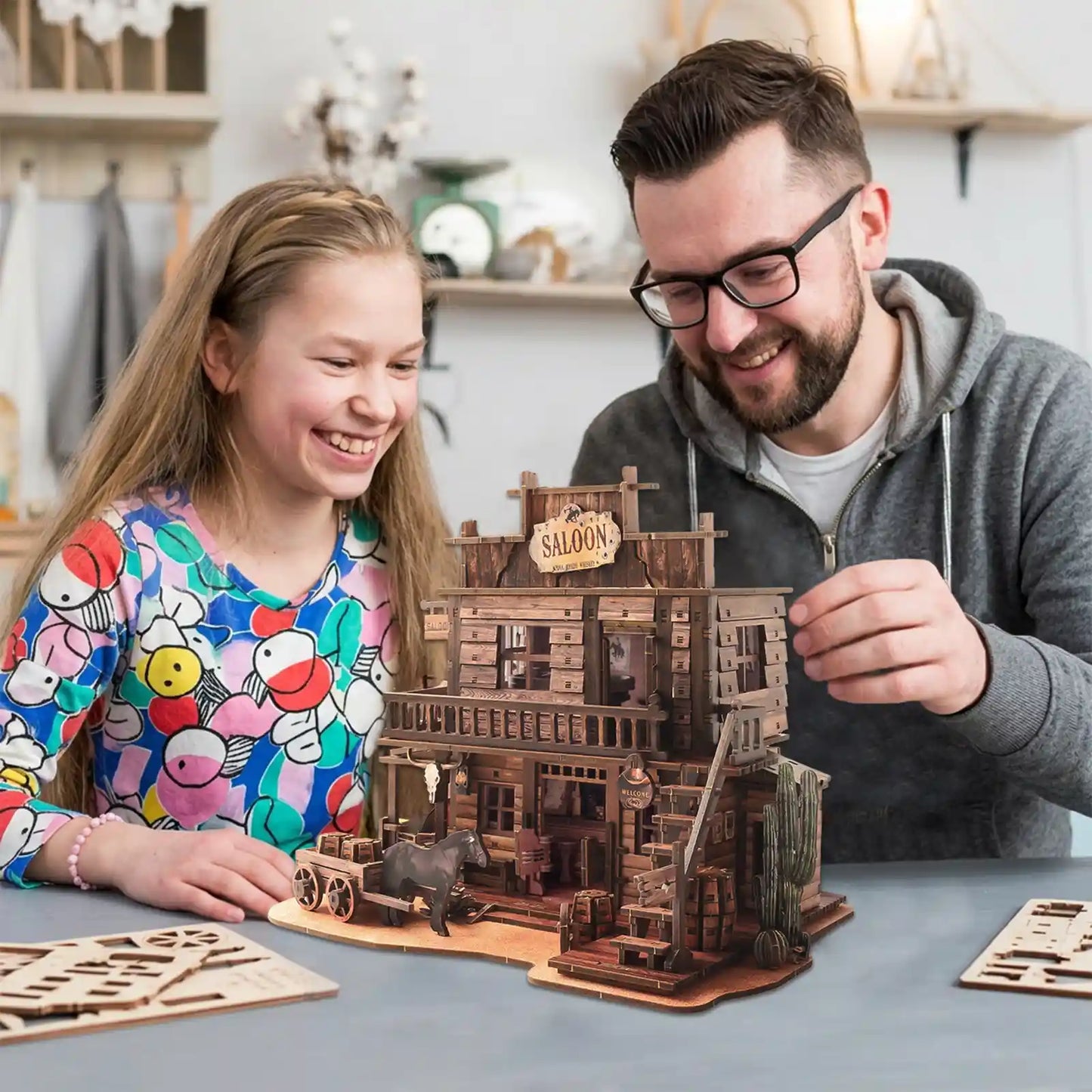 A smiling girl and a man assemble the benlarkytoys Western Saloon DIY Wood 3D Puzzle for Adults, creating a detailed Wild West saloon diorama from laser-cut parts together at a cozy indoor table.