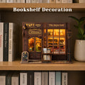 A miniature bookshelf decoration shaped like a cozy bookshop storefront sits on a wooden shelf, surrounded by books and a potted plant. The shop features warm yellow interior lighting, decorative windows, a 'Welcome' sign, and a small streetlamp, with the text 'Bookshelf Decoration' overlaid at the top.