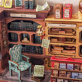 A miniature wooden bookshelf and counter display with toy books, labeled boxes, a small chair, and shelf accessories arranged in a detailed room-like setup.
