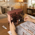A person's hands are assembling a 3D woodenDIY craft kit shaped like a cozy shop storefront, with an instruction booklet and a small tube of adhesive on a wooden table in a warm, living room setting.