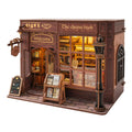 A wooden model of a quaint shop called 'The cheese book' on a clean white background. The miniature building features dark brown wood paneling, warm yellow glowing windows, a street lamp, a sidewalk, and a small sign on the left.
